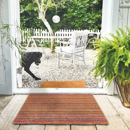 Hug Rug doormat 50cm x 75cm in candy stripe bright pattern placed at entrance of open patio doors overlooking garden with potted fern and black dog on gravel patio