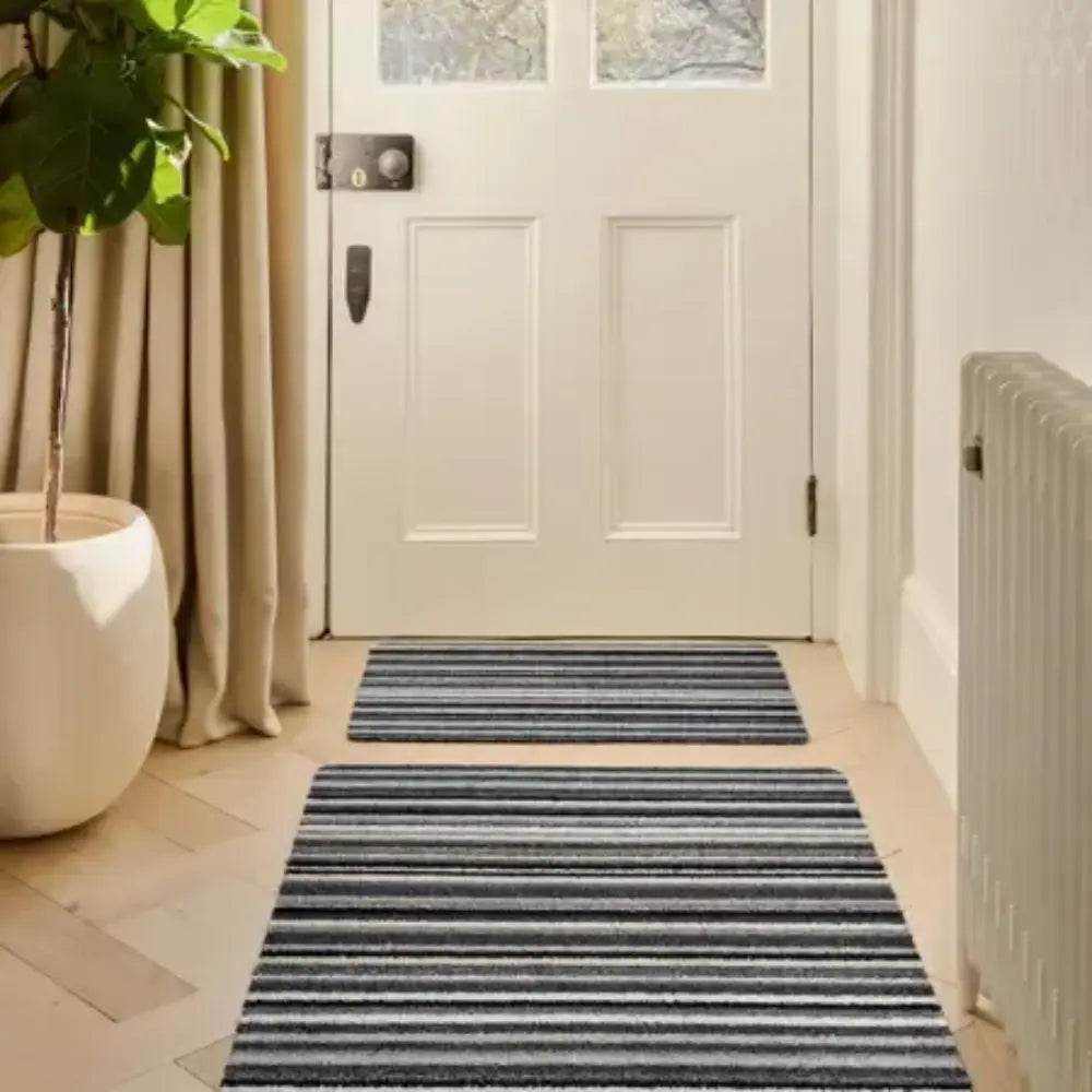 Hug Rug Candy Stripe doormat in charcoal and cream horizontal stripes positioned in a bright hallway entryway with white panelled door, beige walls, and potted plant