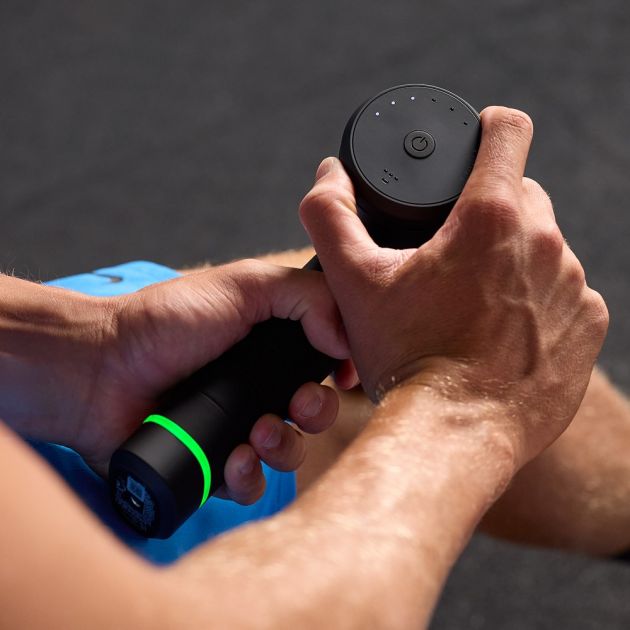 Hands holding black Hyperice Hypervolt 3 Pro massage gun with green accent ring and blue fabric surface visible in background