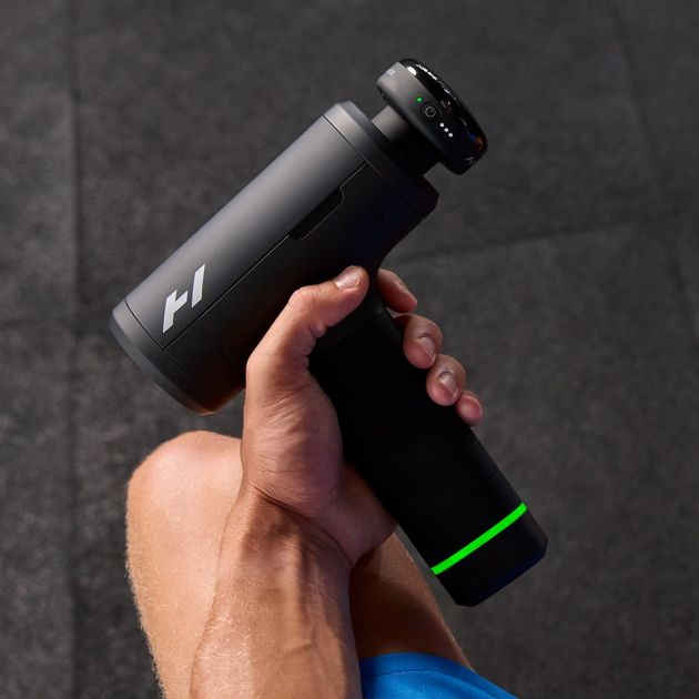 Hand holding Hyperice Hypervolt 3 Pro massage gun in black with green accent stripe and digital display screen against grey background