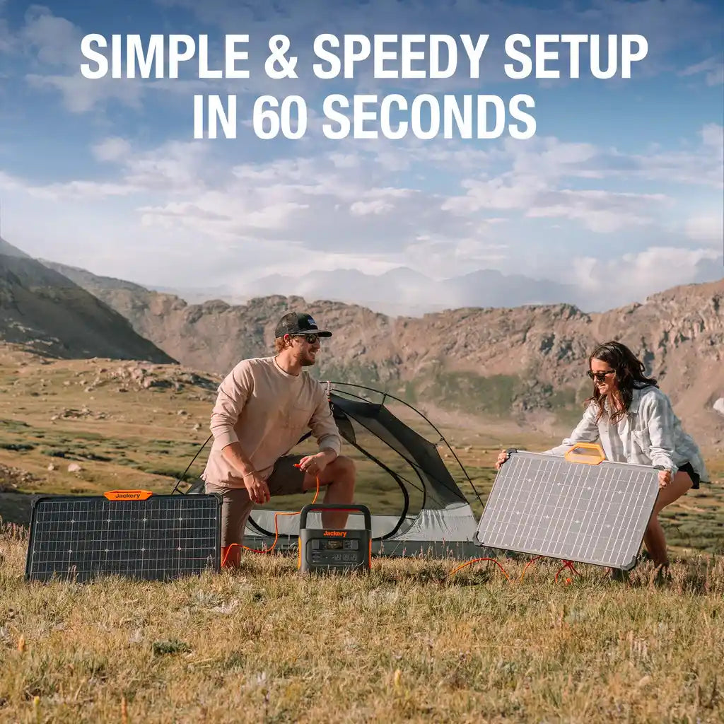 Two people setting up a portable solar power system in a mountainous valley, with a Jackery portable power station connected to multiple foldable solar panels including the SolarSaga 80W panel, demonstrating quick outdoor camping setup