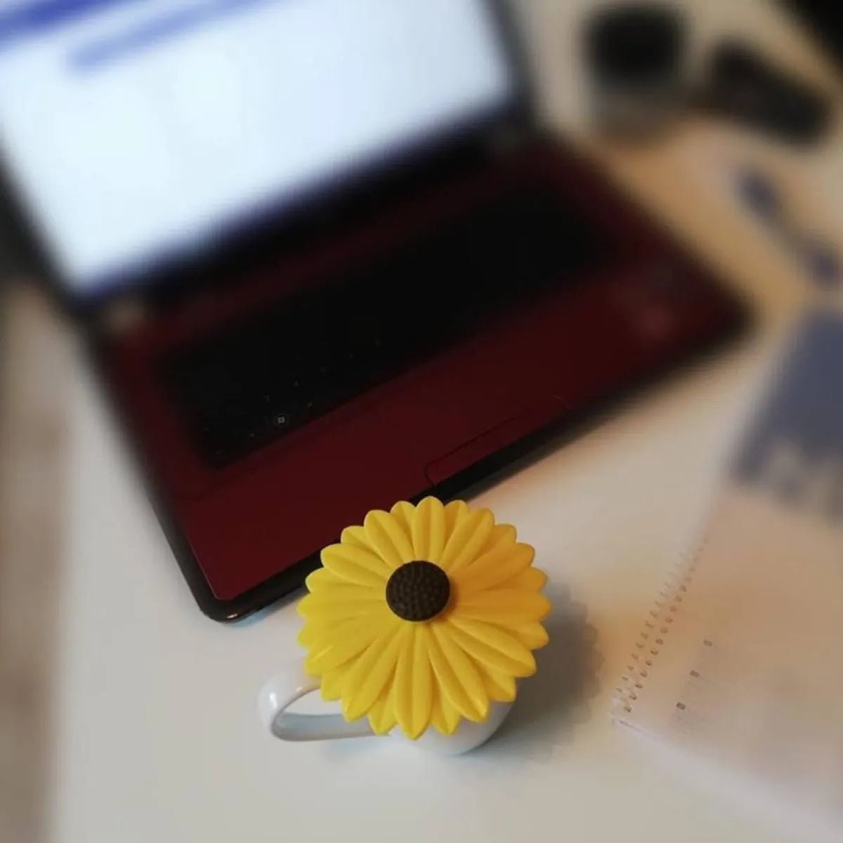 Yellow silicone daisy-shaped cup lid with dark center displayed on a light surface next to a red and black mug, with a smartphone and other items blurred in the background