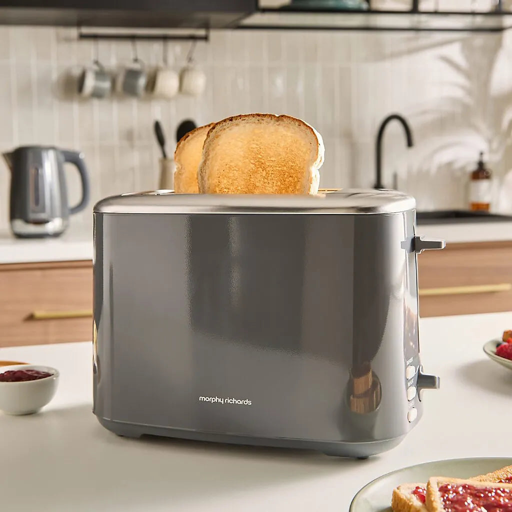 Morphy Richards Equip 2 slice toaster in grey with two slices of toasted bread popping up, displayed on a white kitchen counter with a kettle and breakfast items in the background