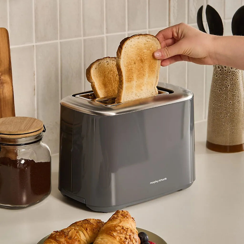 Morphy Richards Equip 2 slice toaster in grey with two slices of golden brown toast being removed, displayed on a white countertop with kitchen accessories including a jar of spread and wooden utensils in the background