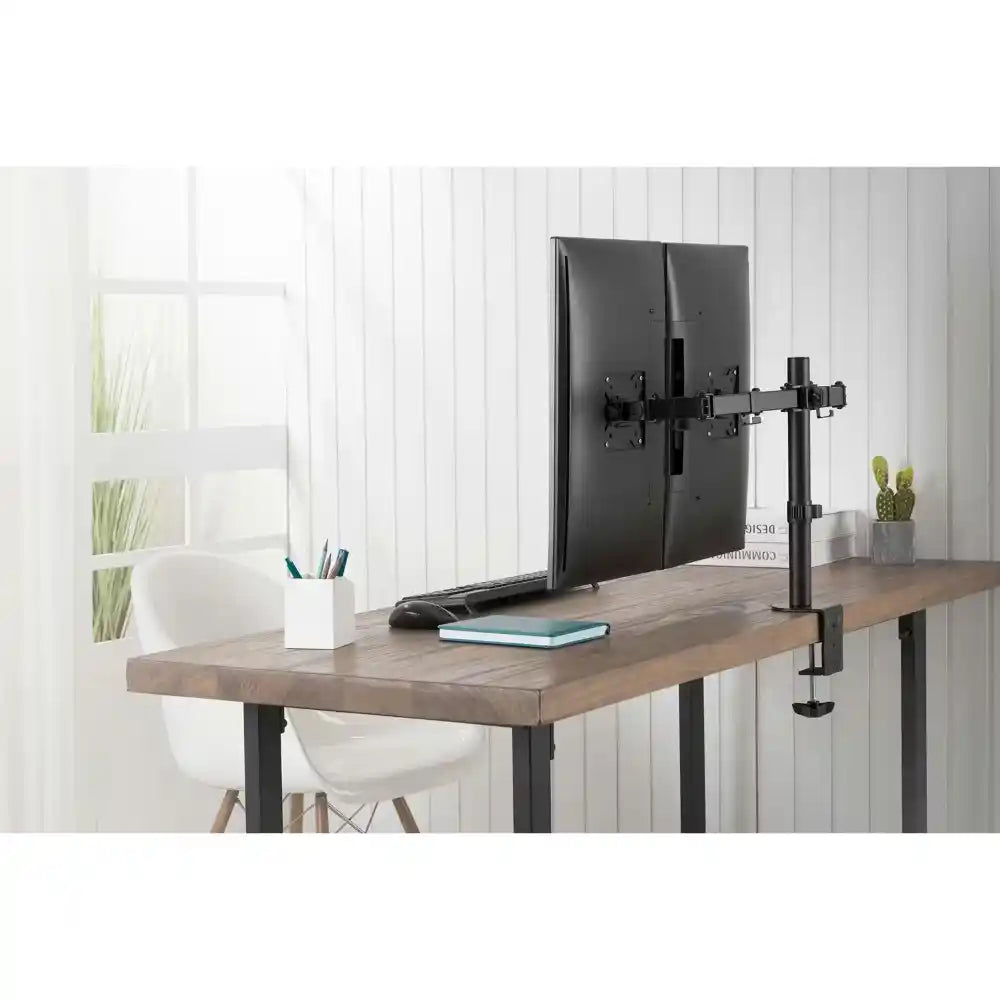 Black dual monitor clamp mount holding two dark monitors on wooden desk with white modern chair, potted plants and desk accessories in bright minimalist office setting