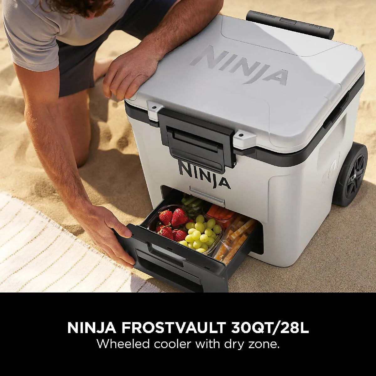 Man opening lower drawer of white and black Ninja FrostVault 28L wheeled cooler displaying fresh strawberries, grapes and snacks inside on sandy beach