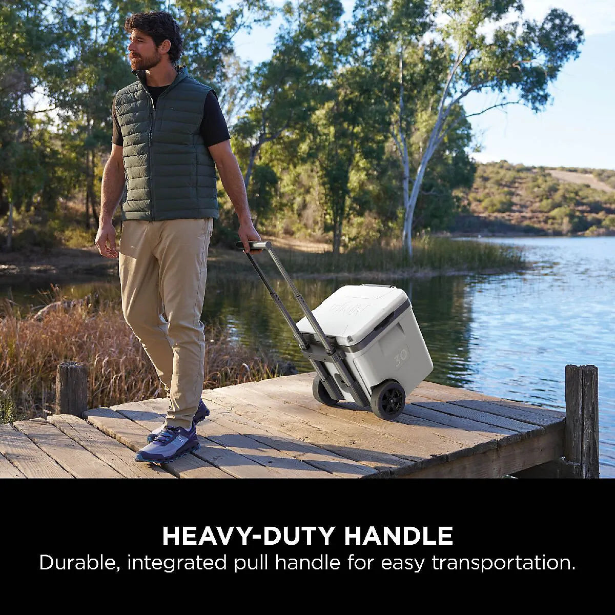 Man in black gilet and beige pants pulling white Ninja FrostVault 28 litre wheeled cooler box with integrated handle on wooden dock beside calm lake surrounded by eucalyptus trees