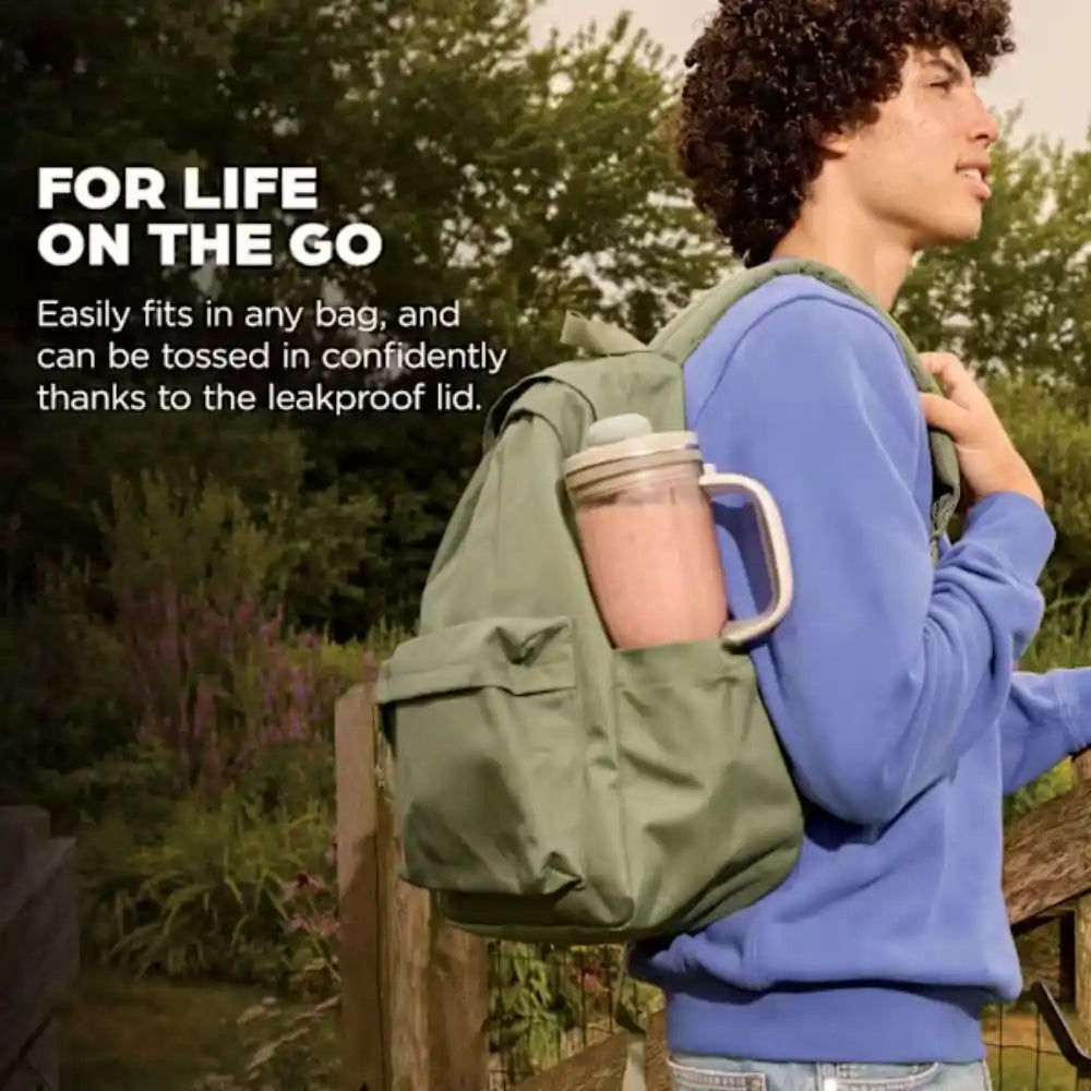 Person wearing blue sweatshirt carrying olive green backpack with pink Ninja BlendBOSS tumbler blender visible, standing outdoors in garden setting with trees and plants in background