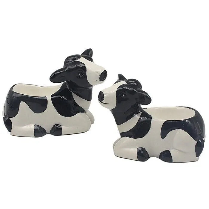 Set of two ceramic cow-shaped egg cups in black and white with glossy finish, featuring detailed facial features and lying down posture, displayed against white background