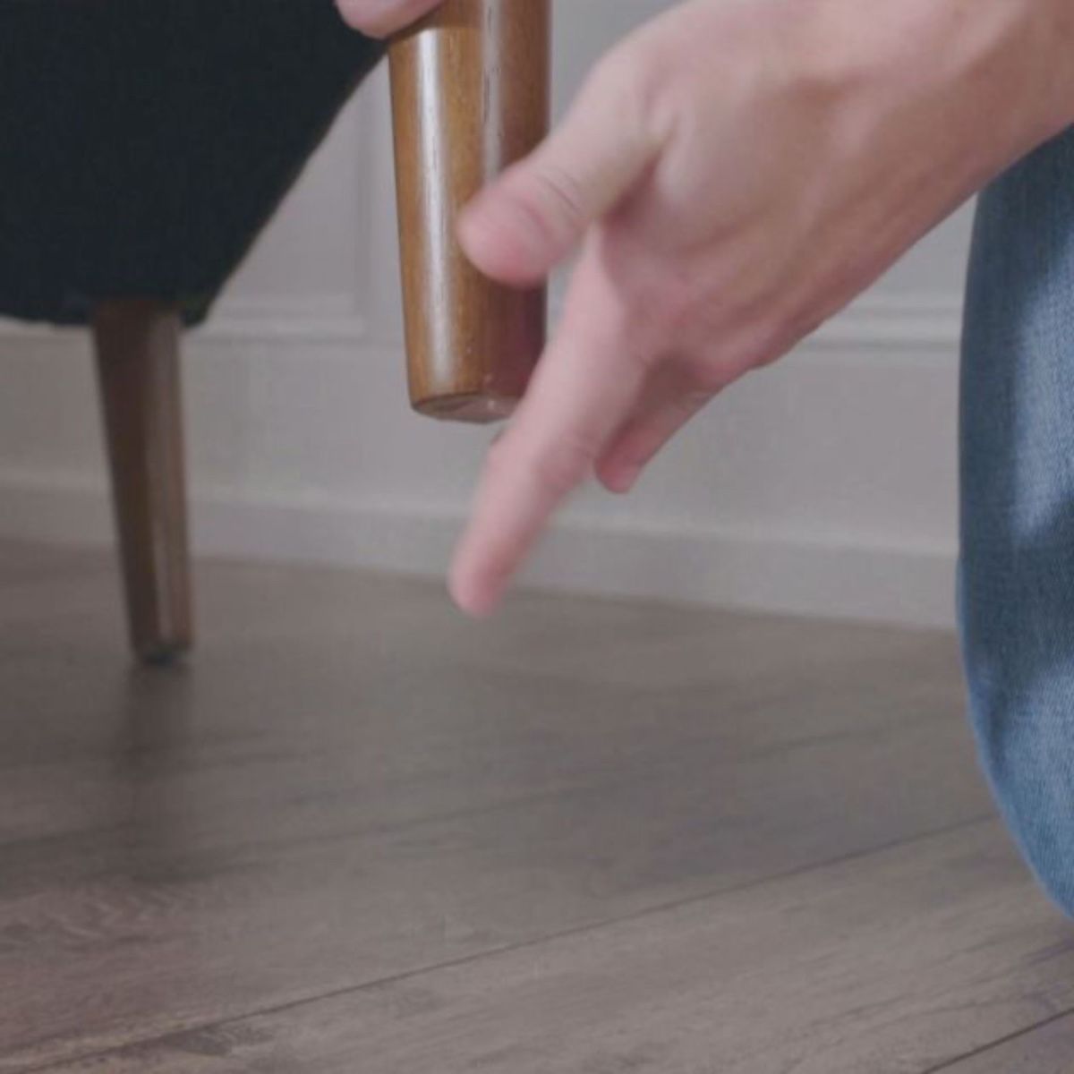 Hand demonstrating JML Starlyf Fantastic double-sided adhesive tape application on wooden furniture leg against hardwood floor, showing tan adhesive tape roll in use