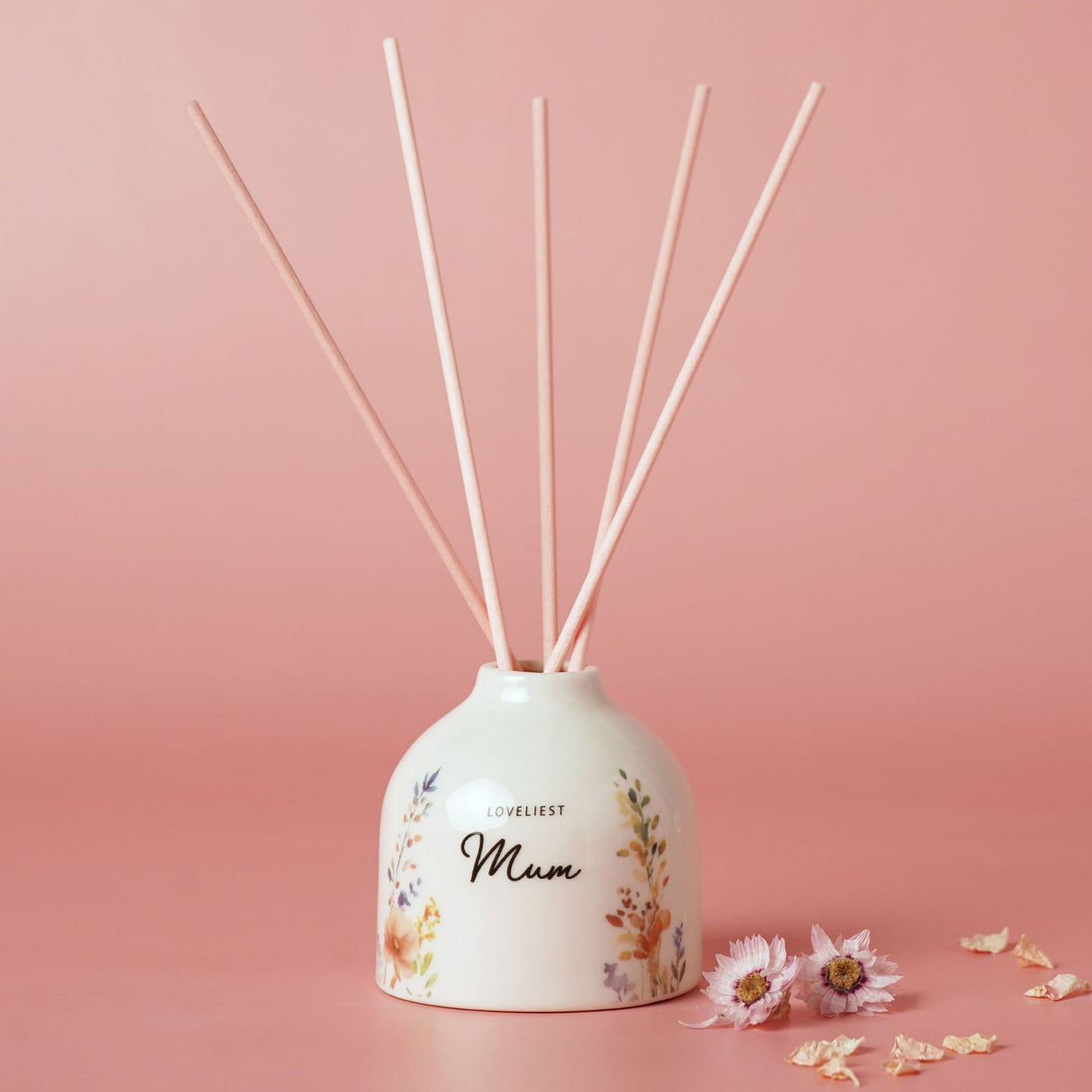Ceramic reed diffuser with "Loveliest Mum" text and watercolor floral design in pink and gold tones, featuring five reed sticks, displayed against a soft pink background with scattered dried flowers
