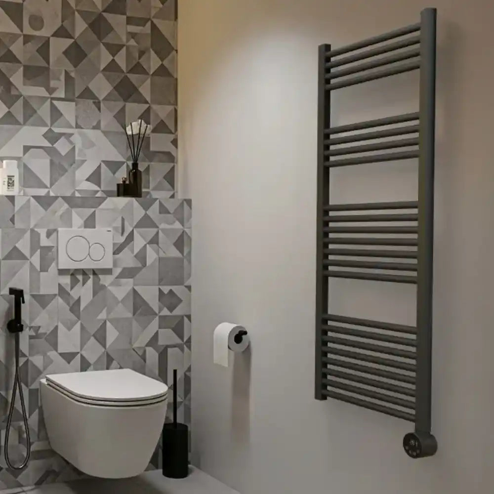Grey heated towel rail with horizontal bars mounted on bathroom wall next to white toilet, featuring modern geometric patterned tiles in grey and white tones with black fixtures and accessories