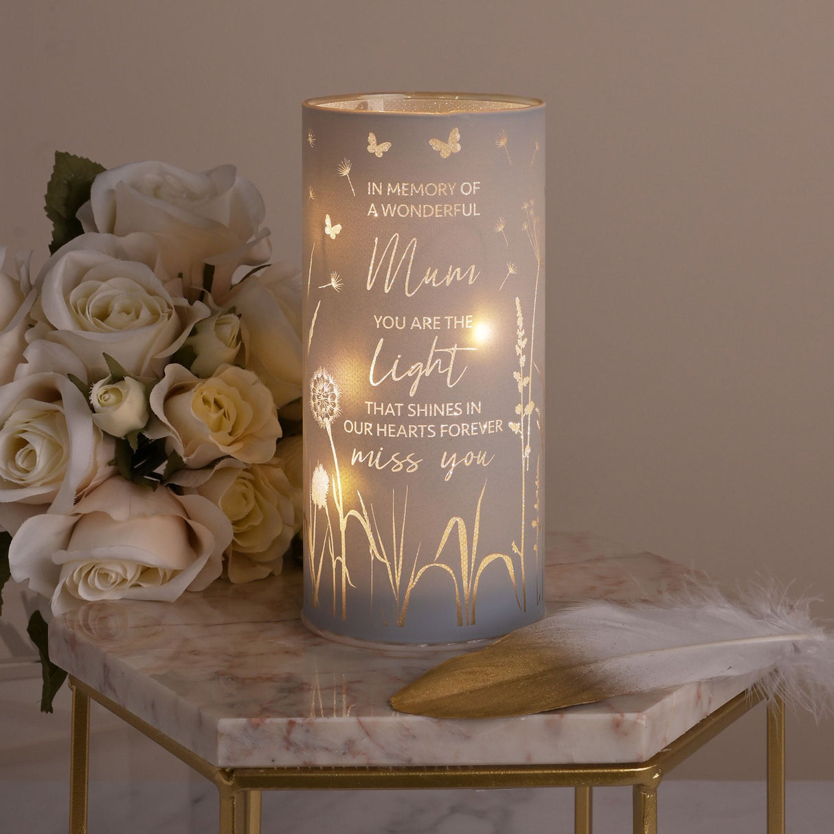 Frosted glass tube light with gold engraved text reading "In Memory of a Wonderful Mum, You Are The Light That Shines in Our Hearts Forever, Miss You" decorated with butterflies and dandelions, illuminated from within, displayed on a marble hexagonal table next to a bouquet of cream and white roses