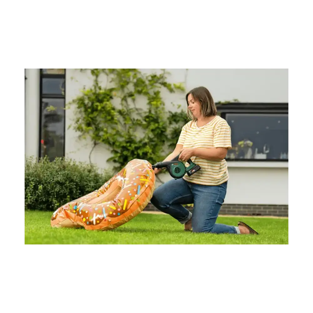 Woman using a Bosch cordless leaf blower on grass in a garden, wearing a yellow and white striped t-shirt and blue jeans, with an inflatable orange dragon toy nearby and a modern house with ivy in the background