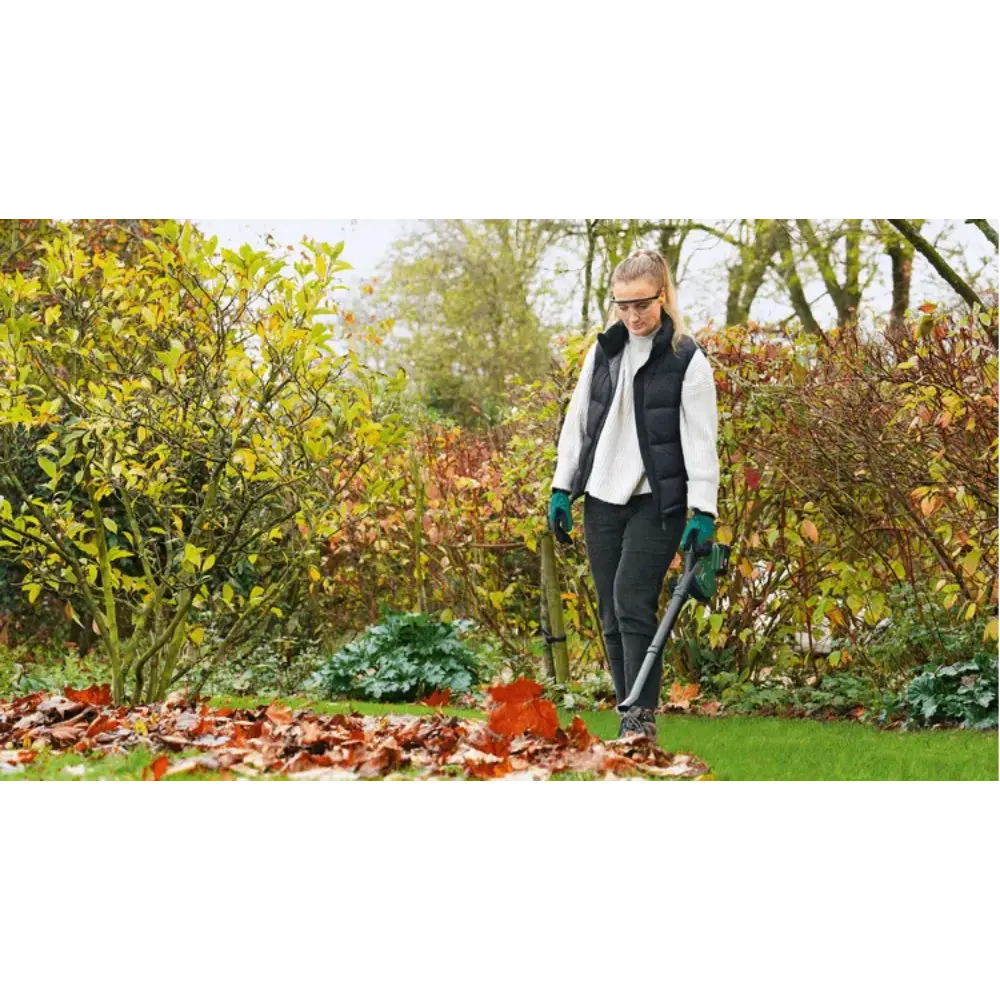 Woman using Bosch Universal Garden Leaf Blower cordless 18V to clear fallen autumn leaves from lawn surrounded by hedges and trees in garden setting