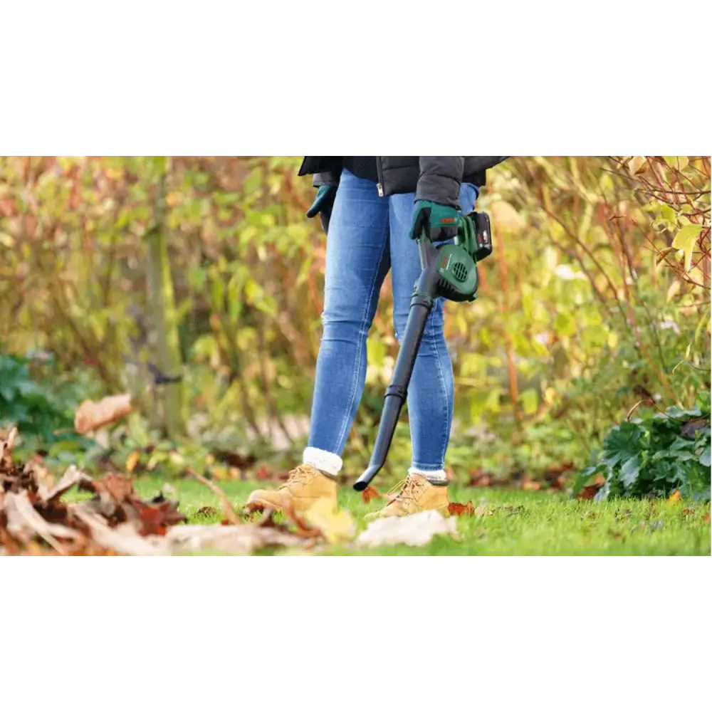 Person wearing blue jeans and tan boots using a Bosch cordless leaf blower with green and black design in an autumn garden surrounded by fallen leaves and shrubs