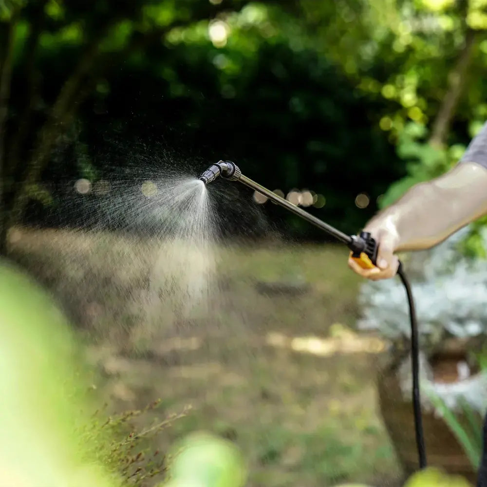 Close-up of WorX WG829E.9 cordless sprayer in action spraying water mist over green garden foliage with blurred background