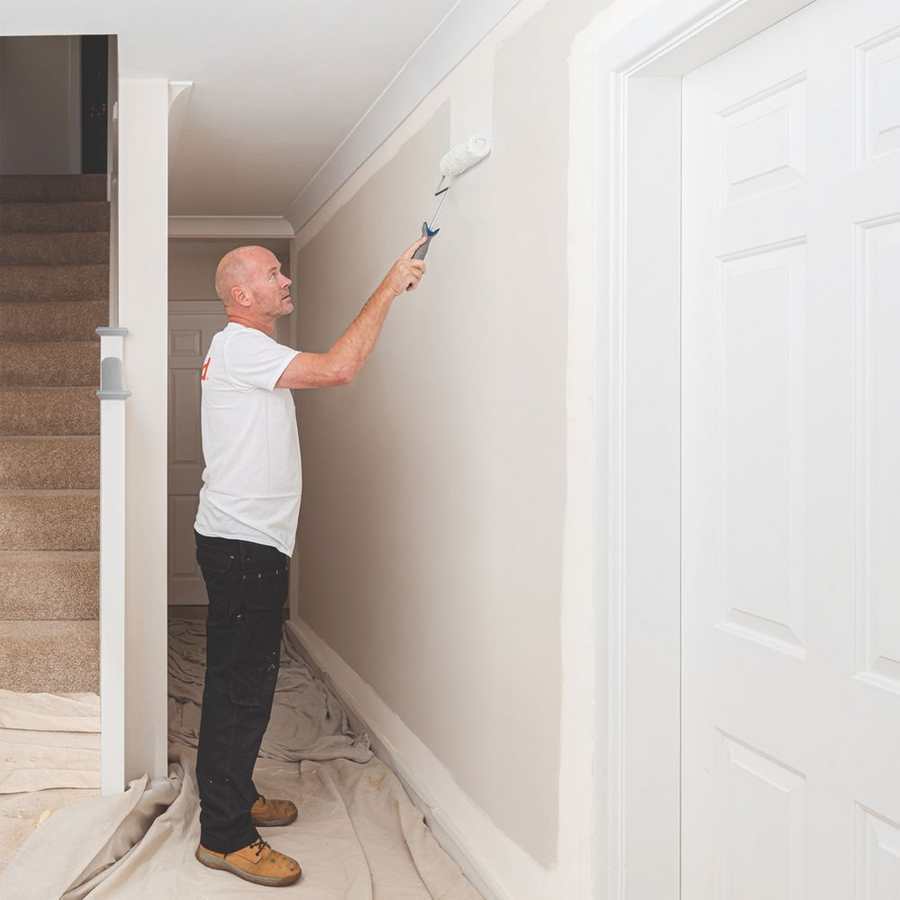 Man painting hallway ceiling with white paint using roller and extension pole, demonstrating Leyland Contract Matt white paint application on residential interior walls and ceiling