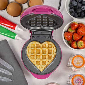 Pink and black heart-shaped waffle maker with golden cooked waffle, surrounded by fresh strawberries, blueberries, eggs, and baking supplies on white surface