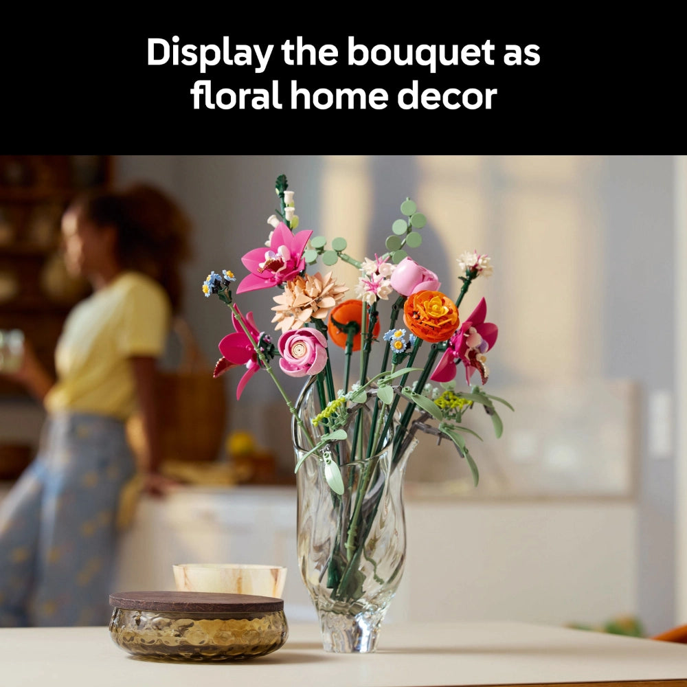 Colorful LEGO flower bouquet displayed in a clear glass vase on a white surface, featuring pink orchids, orange roses, and delicate foliage with green stems, styled as home decor in a bright, modern living room setting