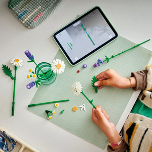 Child's hands assembling green-stemmed LEGO flowers with white and purple petals on a light green mat, with a smartphone displaying a building instruction guide and completed flower pieces arranged nearby