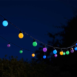 Colorful LED festoon string lights strung across dark evening sky featuring 30 illuminated spherical bulbs in cyan, yellow, green, purple and blue tones against silhouetted trees and foliage