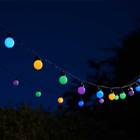 Colorful LED festoon string lights strung across dark evening sky featuring 30 illuminated spherical bulbs in cyan, yellow, green, purple and blue tones against silhouetted trees and foliage