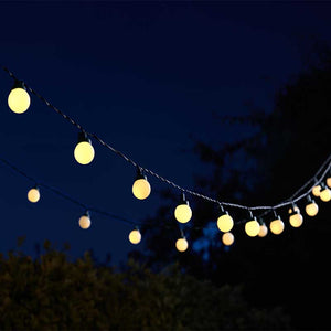 String of 30 warm yellow spherical LED festoon lights draped against a deep blue evening sky with silhouetted trees in the background