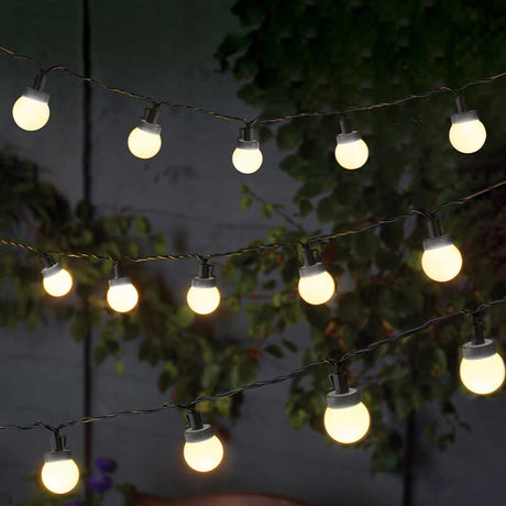 Warm white LED festoon string lights with 30 globe bulbs strung across dark wires, illuminated against an evening garden backdrop with green foliage and grey stone wall