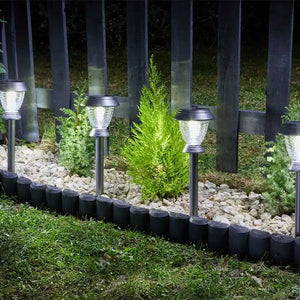 Four stainless steel solar stake lights with clear glass tops arranged in a garden bed among white gravel and green plants, backed by a dark wooden fence, illuminated at night