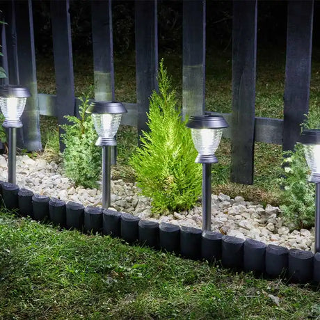 Four stainless steel solar stake lights with clear glass tops arranged in a garden bed among white gravel and green plants, backed by a dark wooden fence, illuminated at night