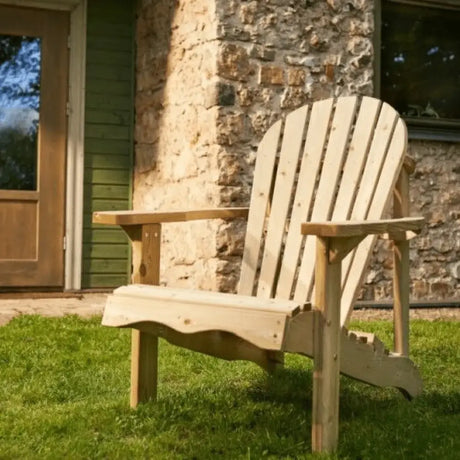 Pressure treated wooden Adirondack garden chair in natural light wood finish positioned on green lawn against stone cottage wall with green door