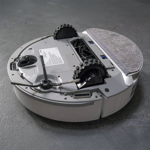 White circular robotic vacuum cleaner with black dual rotating brushes, gray fabric dust collection pad on top, control panel with documentation, power cord, and tread wheels visible from overhead angle on gray surface
