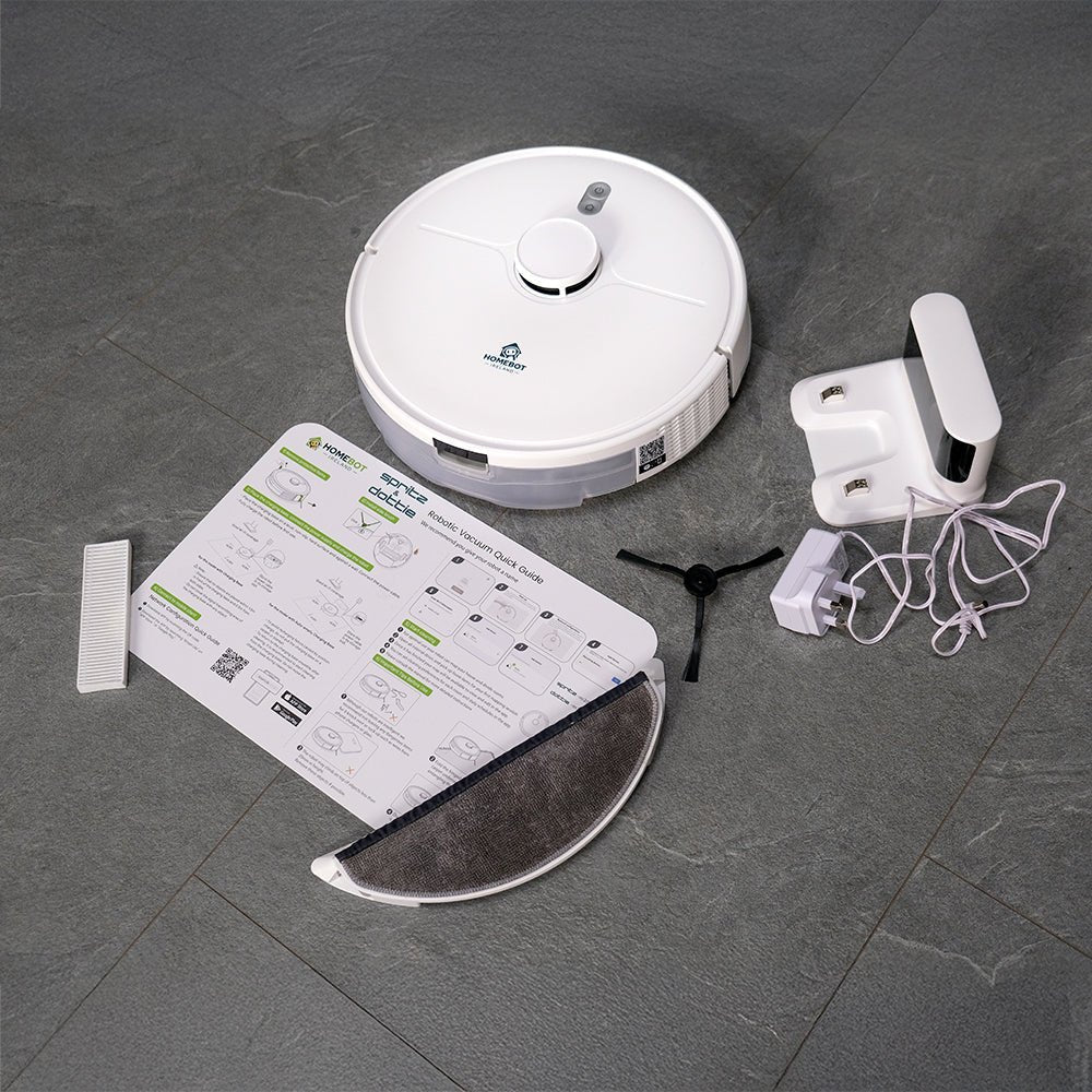 Homebot Spritz robotic vacuum cleaner unboxing flat lay showing white circular robot vacuum, instruction manual with green accents, white charging dock with power cable, and white mopping pad on dark grey tile floor