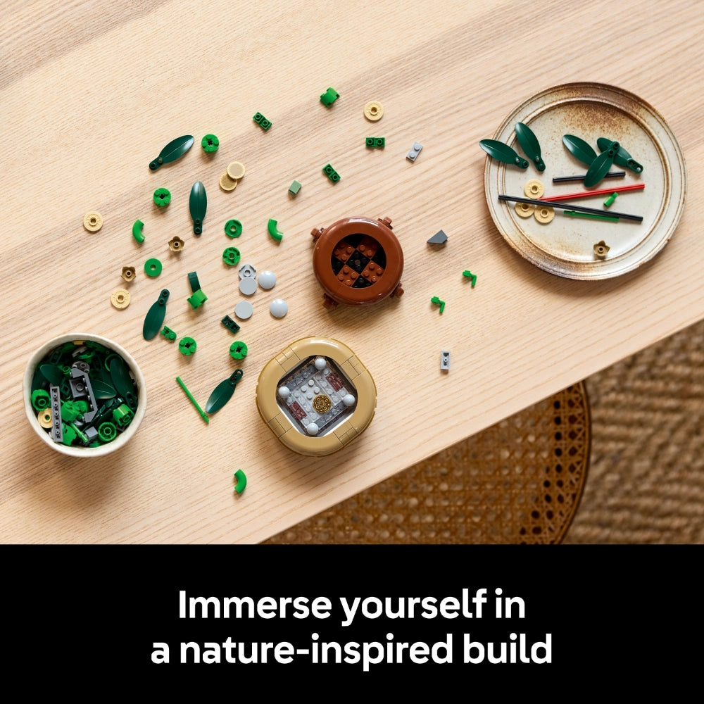 Overhead view of LEGO Lucky Bamboo set 10344 components displayed on light wood surface, featuring green leaf pieces, tan and gold connector pieces, brown ceramic pot, white organizer bowl, and instruction materials arranged in nature-inspired layout