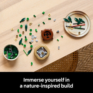 Overhead view of LEGO Lucky Bamboo set 10344 components displayed on light wood surface, featuring green leaf pieces, tan and gold connector pieces, brown ceramic pot, white organizer bowl, and instruction materials arranged in nature-inspired layout