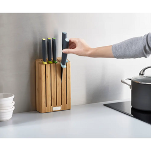 Person removing a kitchen knife with black handle from a wooden bamboo knife block containing five knives with colored accents, displayed on a white countertop next to a black cooking pot and white bowls