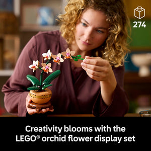 Woman with curly reddish-blonde hair wearing a burgundy shirt examines a LEGO orchid flower display set in a wooden pot with white and pink orchid blooms and green leaves against a blurred home interior background