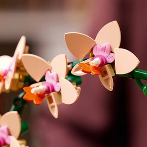 LEGO Mini Orchid 10343 set featuring peach and pink plastic orchid flowers with orange centers, pink stamens, and green stems against a soft blurred background