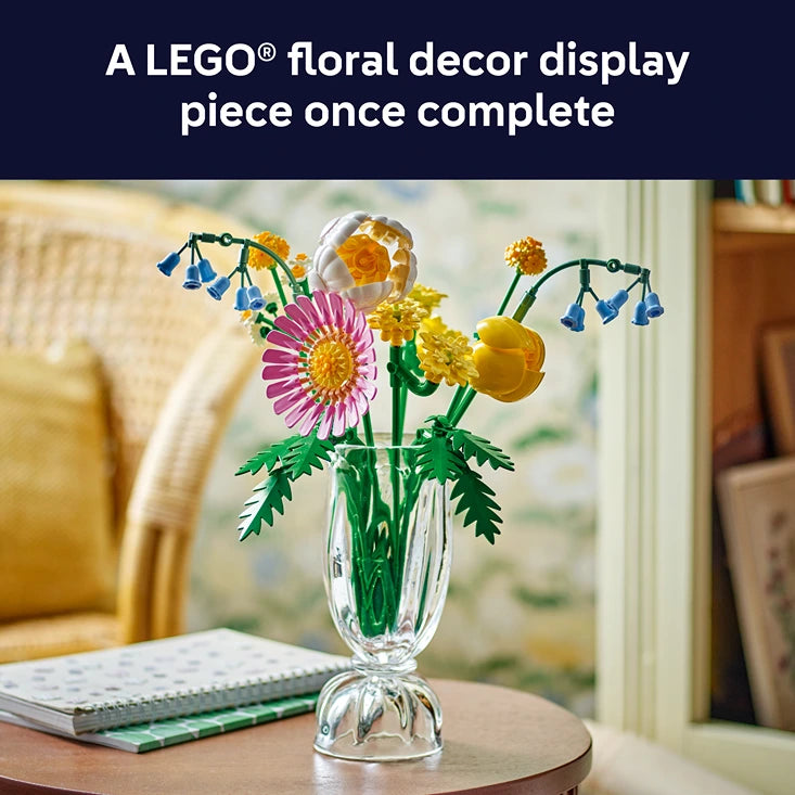 Completed LEGO Petite Sunny Bouquet floral display featuring pink gerbera, yellow daisies, orange ranunculus and blue forget-me-nots with green stems in a clear glass vase, displayed on a wooden table by a window