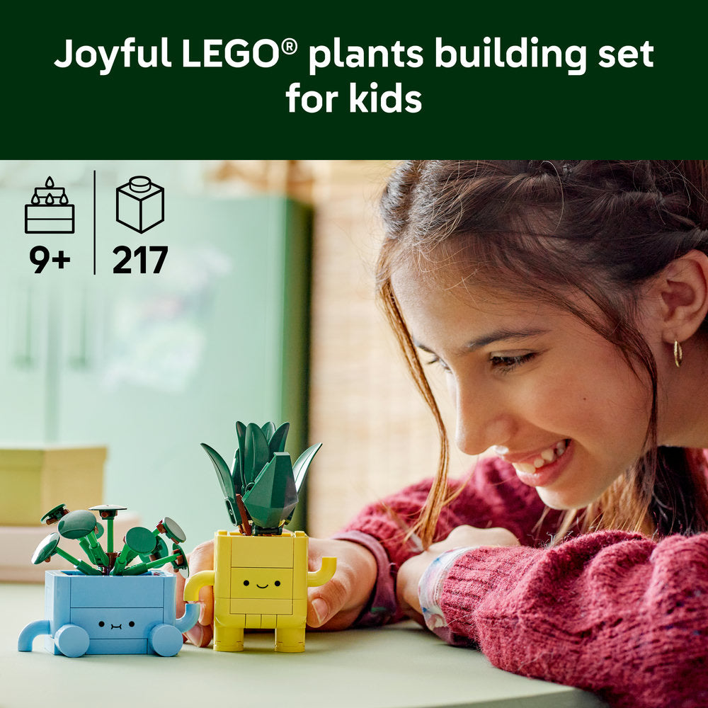 A young girl in a burgundy sweater smiles while playing with Joyful LEGO Plants building set, featuring buildable plant figures in blue and yellow pots with friendly faces, recommended for ages 9+, containing 217 pieces