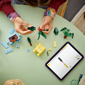Child's hands assembling LEGO Happy Plants set with colorful brick pieces including green plants, yellow smiling character, and blue building blocks on pale green surface, with tablet showing instruction guide
