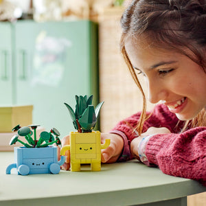 Young child playing with LEGO Happy Plants set featuring a smiling yellow planter pot with green succulent plant and a blue wheeled planter with green foliage on a white table in a bright, colorful room