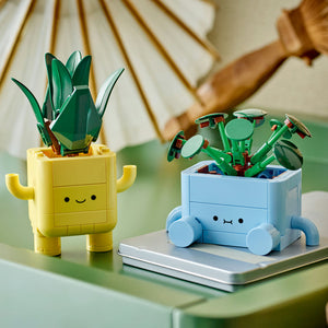 Two LEGO Happy Plants buildable figures displayed on a green surface, featuring a yellow cactus-shaped planter with a smiling face and green succulent leaves, and a blue wheeled planter pot with closed eyes containing a green flowering plant, set against a blurred background with a striped parasol and wooden furniture