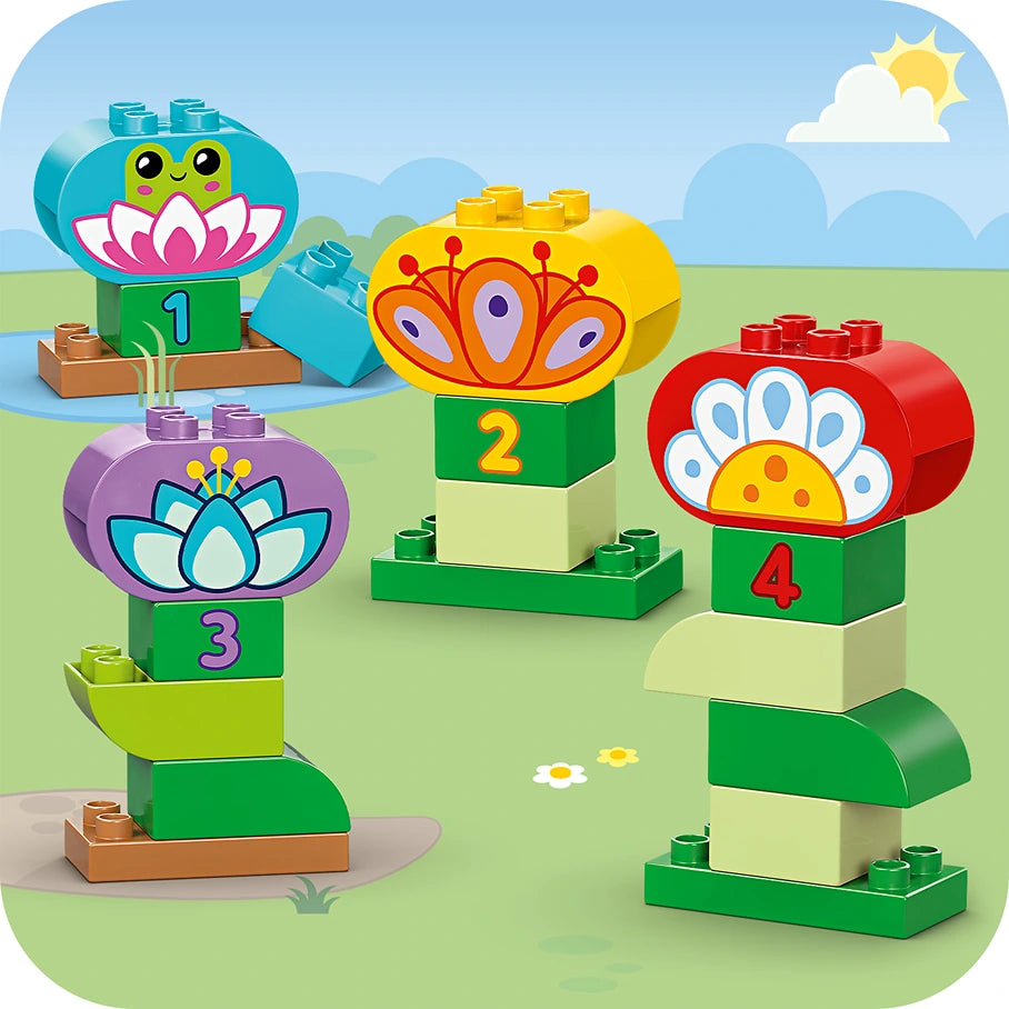 LEGO Duplo Creative Garden and Flowers set 10444 featuring colorful stacked flower-shaped blocks in turquoise, yellow, purple and red with numbered green base blocks, displayed on a bright green grass background with blue sky and sun