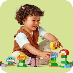 Toddler with curly brown hair playing with colorful LEGO Creative Garden and Flowers building set on yellow background, featuring green base plates, flower-themed blocks with numbered pieces, and bee-shaped elements