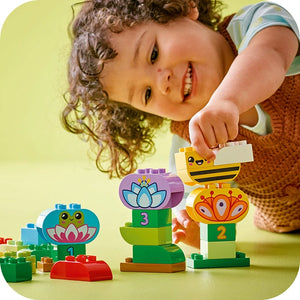 Toddler with curly hair playing with LEGO Duplo Creative Garden and Flowers set 10444, featuring colorful stacked blocks with flower and bee designs in purple, green, yellow and orange against a yellow background
