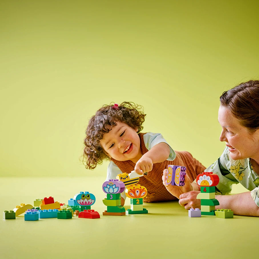 Adult and young child playing together with colorful LEGO Creative Garden and Flowers building set on a bright yellow background, featuring stacked flower-shaped bricks with purple, yellow, orange and red details, green stems and leaves, demonstrating creative play and construction