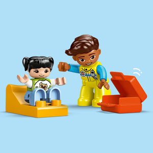 LEGO Duplo ambulance set featuring two smiling figurines, a yellow ambulance vehicle with blue and white details, and a red emergency medical box against a light blue background