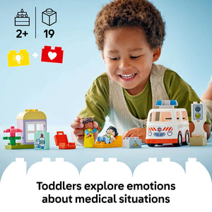 A young child with curly brown hair wearing a green shirt plays with a LEGO Duplo ambulance set featuring colorful building blocks, minifigures, and a white and orange ambulance vehicle. The image shows age recommendation 2+ and 19 pieces, with icons indicating it supports emotional learning about medical situations. Light blue background with displayed set components including a small house, figures, and medical-themed accessories